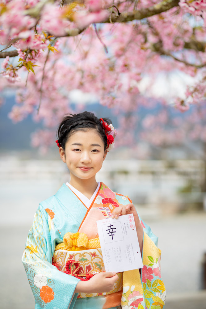 想いを込めた一文字を書いた紙を持って、桜の前で笑顔でポーズをとる女の子の写真