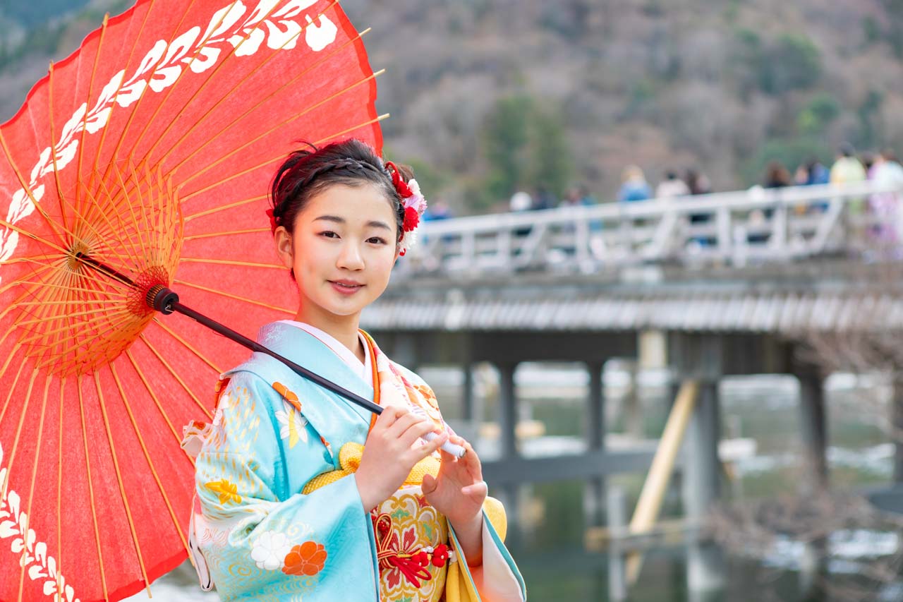 嵐山の渡月橋を背景に赤い和傘を持つ、着物を着た少女の写真